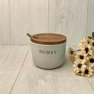 May include: A white ceramic honey pot with a wooden lid and a small wooden spoon. The word "HONEY" is printed on the front of the pot in a dark gray font. A bouquet of white and brown flowers is to the right.