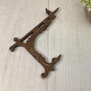 May include: Ornate bronze-colored picture frame stand with a decorative floral design. The stand has a curved base and a hinged top, designed to hold a picture or artwork. The stand is made of metal and has a vintage aesthetic.