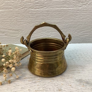 May include: A small, brass, decorative pot with a handle. The pot has a rounded bottom and a flared top. The handle is ornate and has a curved shape.