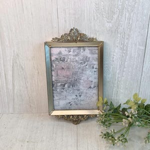 May include: A rectangular picture frame with a metallic gold finish and ornate, decorative scrollwork detailing at the top and bottom edges. The frame contains a printed insert featuring a soft, muted floral pattern in shades of gray and pale pink.