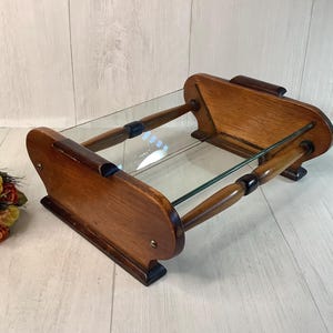 May include: A vintage serving tray with a clear glass surface and a wooden frame. The tray has a rectangular shape with rounded ends and is supported by wooden legs. The wood has a warm, brown tone, and the tray appears to be in good condition.