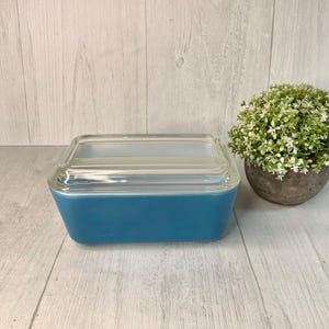 May include: A rectangular, turquoise-colored Pyrex dish with a clear glass lid. The dish is on a light wood-grain surface, next to a small potted plant with green and white foliage. The dish is a vintage kitchen item.