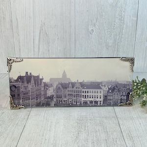 May include: A rectangular framed print featuring a black and white cityscape with ornate silver corner accents. The image depicts buildings and a church steeple. The frame is resting on a light wood surface.
