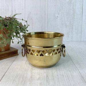 May include: A polished brass planter featuring a decorative band of cut-out heart shapes around the upper section. The round container includes two side handles with attached rings and a smooth, flared rim, presenting a metallic gold finish.