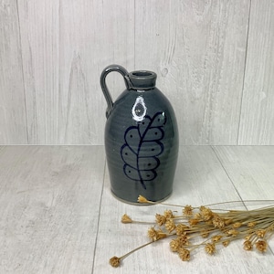 May include: A dark gray ceramic jug with a handle and a dark blue leaf design. The jug is oval-shaped with a narrow neck. Dried flowers are in the foreground. The jug is likely used for decorative purposes.