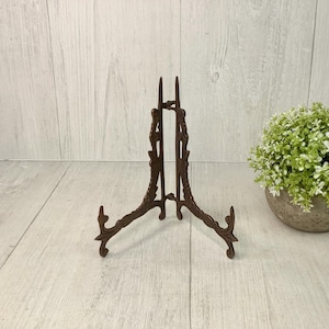 May include: A decorative, brown metal plate stand with an ornate, scrolled design. The three-legged stand is designed to hold a plate or artwork. A small, round, potted plant with green and white foliage is to the right.