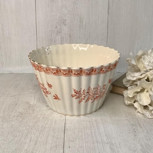 May include: Cream-colored ceramic bowl with a fluted design and a decorative orange floral pattern around the rim and on the sides. The bowl has a scalloped edge and is likely vintage or antique.