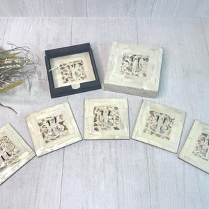 May include: A set of five square coasters and a matching square storage box, all featuring a white mother-of-pearl inlay border surrounding a central mosaic of assorted natural seashells and fragments set in a light-colored resin material.