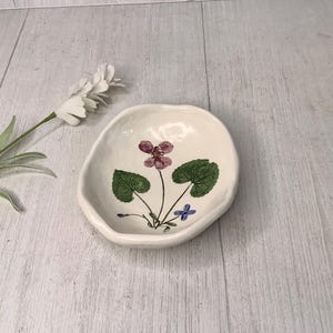 May include: Off-white ceramic dish with a floral design featuring a purple flower, green leaves, and blue accents. The dish has an irregular shape. A white flower and green stem are in the background.