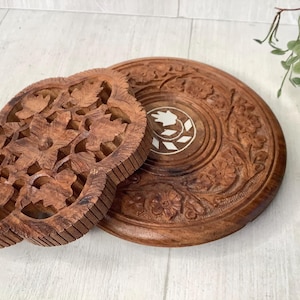 May include: Two intricately carved wooden coasters. One is round with floral patterns and a white inlay. The other is a four-leaf clover shape with detailed leaf and flower carvings. Both are made of a warm brown wood.