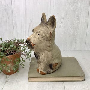 May include: A vintage dog sculpture in a seated position, painted in shades of gray and brown, with visible wear and tear. The sculpture is placed on a weathered book. A small potted plant with green foliage sits to the left.