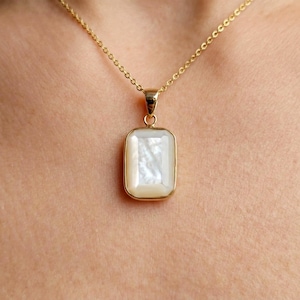 May include: A gold-colored necklace with a rectangular pendant. The pendant features a white, iridescent stone set in a gold frame. The necklace is displayed against a skin-toned background.