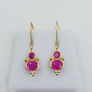Könnte beinhalten: Ein Paar goldfarbene Ohrhänger. Jeder Ohrring hat einen ovalen, magenta-farbenen Edelstein unten und einen kleineren, runden, magenta-farbenen Edelstein darüber. Die Ohrringe haben ein dekoratives Design mit kleinen goldenen Perlen.