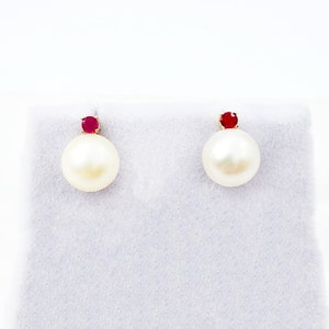 Peut inclure: Une paire de boucles d'oreilles en perles avec une petite pierre précieuse rouge et ronde sur le dessus. Les boucles d'oreilles sont présentées sur une surface blanche. Les perles sont grandes et rondes, de couleur blanc crème.