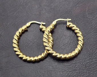 May include: A pair of gold-colored hoop earrings with a twisted rope design. The earrings are made of a shiny, reflective metal and are displayed against a dark background. The hoops have a classic, elegant style.