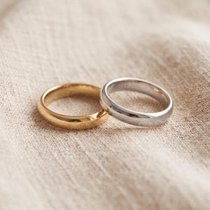 May include: Two smooth, polished metal rings are positioned side by side. One ring is a warm, yellow gold tone, and the other is a cool, silver tone. Both bands feature a simple, rounded profile with a reflective surface and a plain, unadorned design.