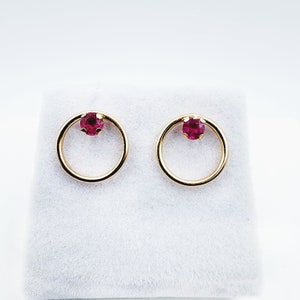Peut inclure: Une paire de boucles d'oreilles créoles en or avec une seule pierre précieuse rose au centre de chaque créole.
