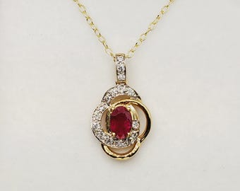 Op de afbeelding: Een goudkleurige ketting met een hanger met een ovale robijn omringd door kleine diamanten. De hanger heeft een bloemmotief met een fijne ketting. De robijn is dieprood en de diamanten schitteren.