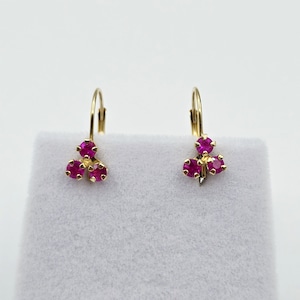 May include: A pair of gold-colored leverback earrings. Each earring features three round, vibrant red gemstones clustered together. The earrings are displayed against a white background, highlighting their design and color.