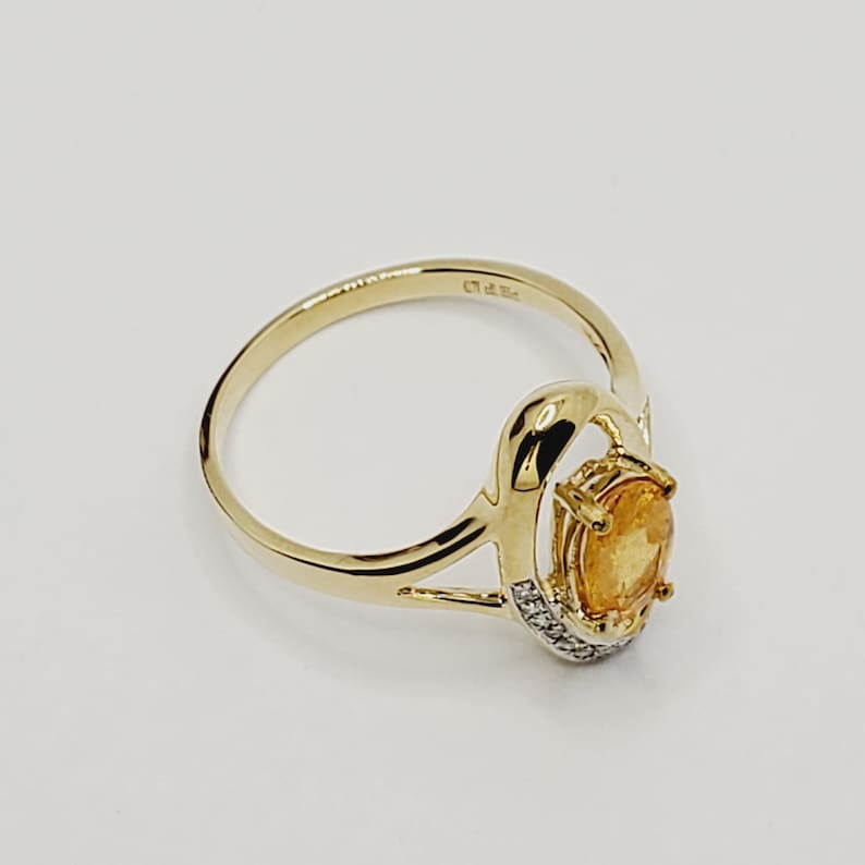 May include: A gold ring with a large oval-shaped orange gemstone surrounded by smaller diamonds. The ring has a unique design with a curved band and a split shank.