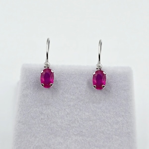 May include: A pair of silver drop earrings with oval, deep pink gemstones. Each earring has a small, round accent stone above the pink gem. The earrings are displayed on a light grey surface against a white background.