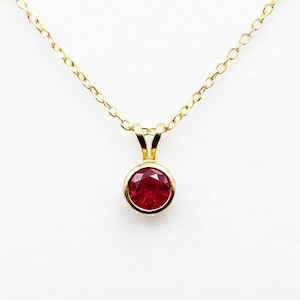May include: A gold necklace featuring a round, faceted red ruby gemstone set in a simple gold bezel pendant. The pendant is suspended from a delicate gold chain with small, uniform oval links, creating a classic and refined jewelry piece.