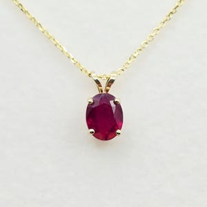 May include: A gold chain necklace featuring an oval-cut deep red ruby pendant held by four prongs. The gemstone is set in a gold mounting that connects to a delicate gold link chain, creating a simple and elegant piece of jewelry.