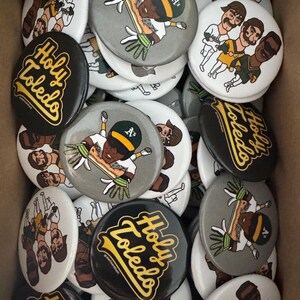 May include: A collection of round buttons with various designs. Some buttons feature cartoon illustrations of baseball players in green and gold uniforms. Other buttons display the text "Holy Toledo" in yellow script on a black background.