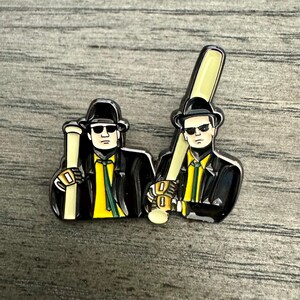 May include: Two enamel pins of men wearing black suits and sunglasses, holding baseball bats. The men are wearing yellow ties and have black hats. The pins are on a wood background.