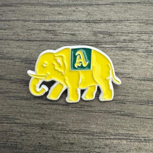 May include: A yellow elephant pin with a white outline. The pin features a green square with a white letter "A" on its side. The pin is on a wooden surface.