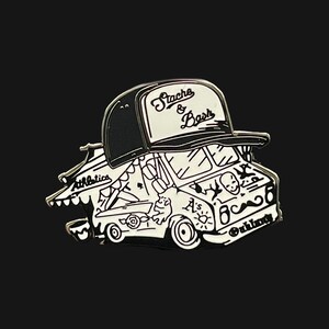 May include: Black and white enamel pin of a food truck with a black and white baseball cap on top. The truck has a mustache and the text "Stache & Bash" on the cap. The truck has the text "Athletics" on the side and "Outlaw" on the back.