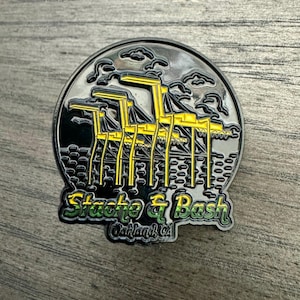 May include: A black and yellow enamel pin with a depiction of a harbor scene with cranes and the text "Stache & Bash Oakland, CA".