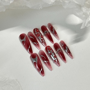 Puede incluir: Conjunto de diez uñas postizas largas, rojas, con degradado, con remolinos plateados y joyas rojas.