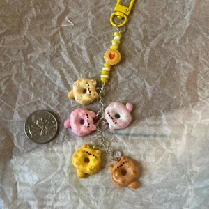 K&ouml;nnte beinhalten: Ein gelber Schl&uuml;sselanh&auml;nger mit einem Donut-f&ouml;rmigen Anh&auml;nger mit einem Katzenkopf. Der Schl&uuml;sselanh&auml;nger hat eine Vielzahl von Donut-Anh&auml;ngern in verschiedenen Farben, darunter rosa, gelb und braun. Die Anh&auml;nger sind an einer silbernen Kette mit einer gelben Perle befestigt.