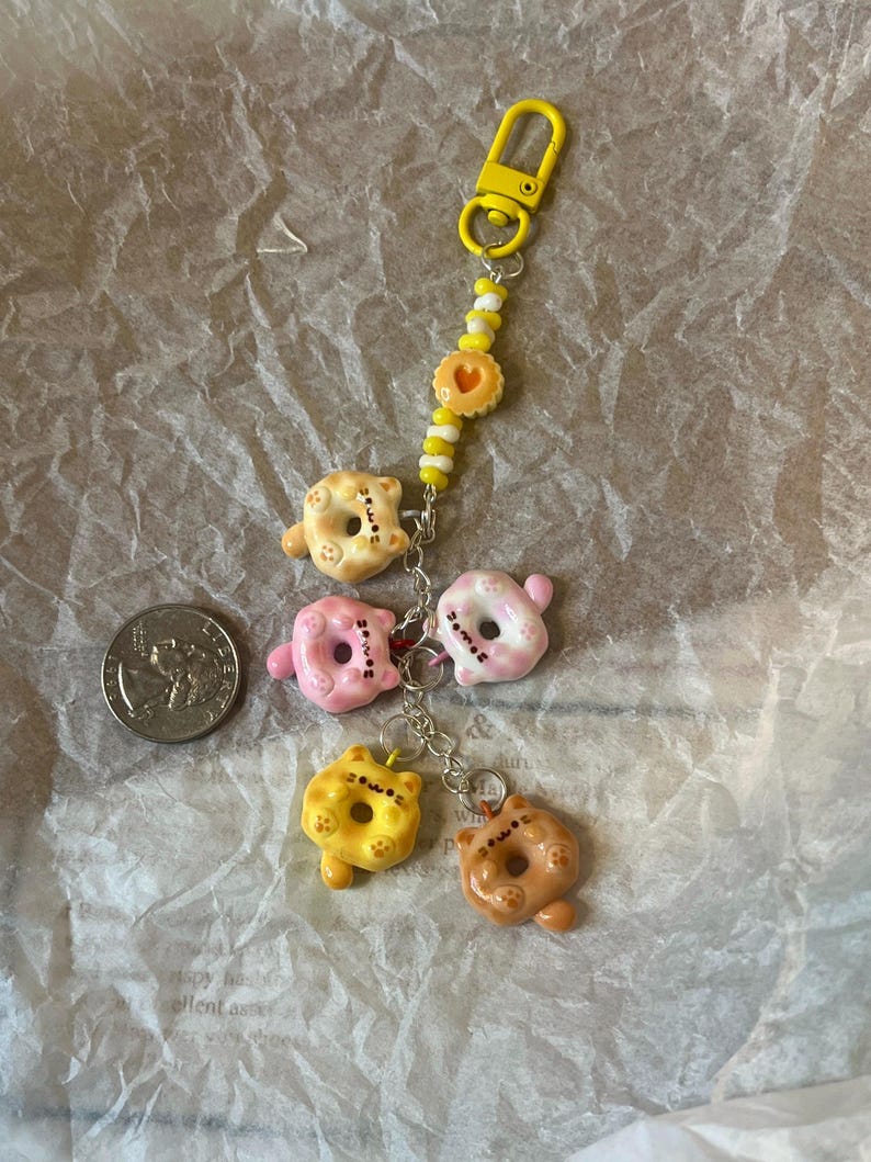K&ouml;nnte beinhalten: Ein gelber Schl&uuml;sselanh&auml;nger mit einem Donut-f&ouml;rmigen Anh&auml;nger mit einem Katzenkopf. Der Schl&uuml;sselanh&auml;nger hat eine Vielzahl von Donut-Anh&auml;ngern in verschiedenen Farben, darunter rosa, gelb und braun. Die Anh&auml;nger sind an einer silbernen Kette mit einer gelben Perle befestigt.