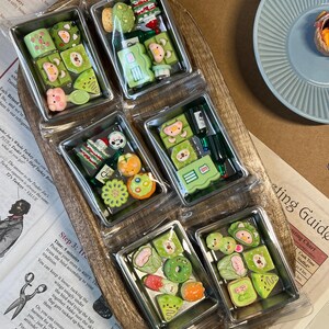 May include: A wooden tray holds six clear plastic containers, each filled with miniature, colorful, food-themed polymer clay creations. The designs include sushi, fruit, and other whimsical shapes, perfect for crafting or collecting.
