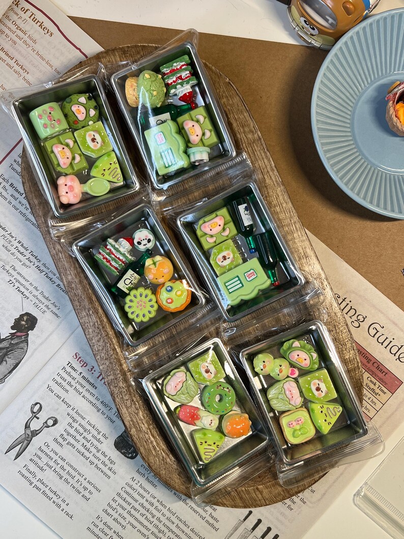 May include: A wooden tray holds six clear plastic containers, each filled with miniature, colorful, food-themed polymer clay creations. The designs include sushi, fruit, and other whimsical shapes, perfect for crafting or collecting.