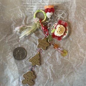 K&ouml;nnte beinhalten: Ein farbenfrohes Charm-Armband mit einem Lebkuchenmann, einem Weihnachtsbaum, einem Stern mit der Aufschrift "Beste W&uuml;nsche" und einem Teller mit einem b&auml;renf&ouml;rmigen Kuchen. Das Armband enth&auml;lt auch ein rot-wei&szlig;es Karomuster, einen gr&uuml;nen Ring und einen rot-wei&szlig;en Erdbeerkuchen.
