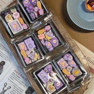 May include: A collection of miniature, decorative resin charms in various shapes and colors, including bears, houses, and slippers. The charms are primarily purple, pink, and yellow, packaged in clear plastic containers.
