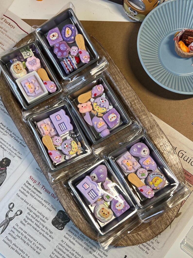 May include: A collection of miniature, decorative resin charms in various shapes and colors, including bears, houses, and slippers. The charms are primarily purple, pink, and yellow, packaged in clear plastic containers.