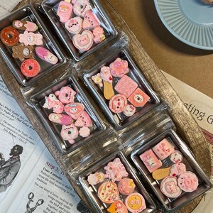 May include: Six clear plastic containers filled with various pink and white miniature food-themed charms. The charms include donuts, bears, and other cute designs. The containers are arranged on a wooden tray.