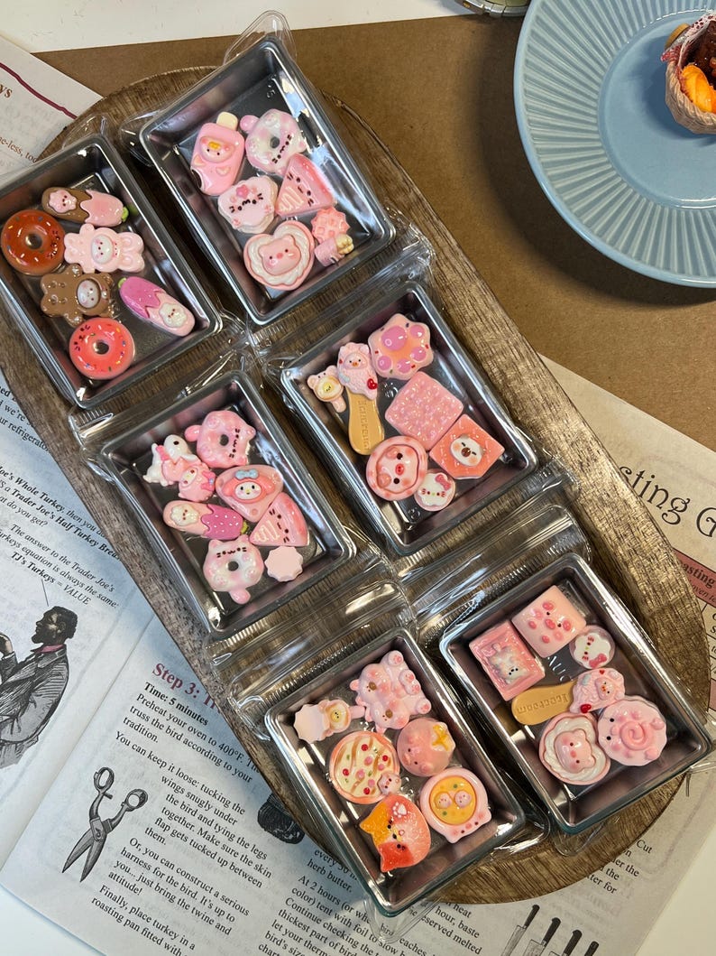 May include: Six clear plastic containers filled with various pink and white miniature food-themed charms. The charms include donuts, bears, and other cute designs. The containers are arranged on a wooden tray.