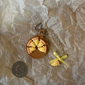 K&ouml;nnte beinhalten: Ein Schl&uuml;sselanh&auml;nger mit einer Miniaturpizza auf einem braunen Holzbrett mit einem gelben Band und einem einzelnen Pizzast&uuml;ck-Anh&auml;nger.