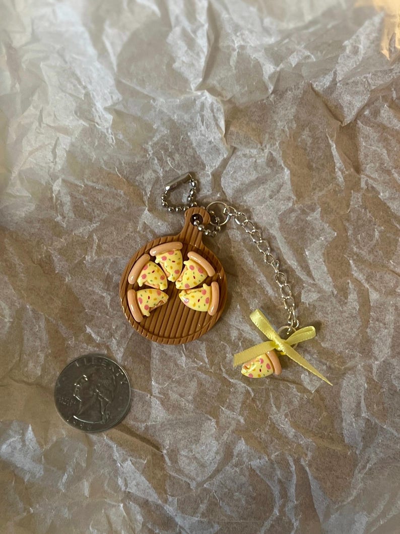 K&ouml;nnte beinhalten: Ein Schl&uuml;sselanh&auml;nger mit einer Miniaturpizza auf einem braunen Holzbrett mit einem gelben Band und einem einzelnen Pizzast&uuml;ck-Anh&auml;nger.