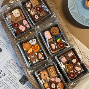 May include: Miniature food charms in clear plastic containers. Each container holds several small, colorful, and detailed food-themed charms, including bears, cookies, and donuts. The charms are arranged on a wooden tray.