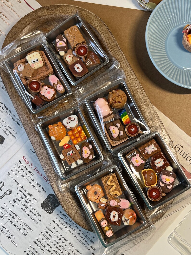 May include: Miniature food charms in clear plastic containers. Each container holds several small, colorful, and detailed food-themed charms, including bears, cookies, and donuts. The charms are arranged on a wooden tray.