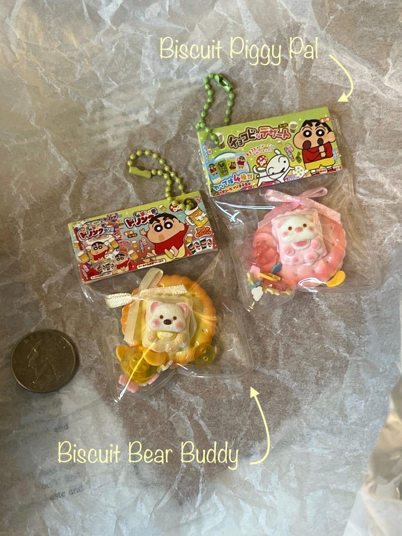 K&ouml;nnte beinhalten: Zwei kleine, bunte Plastik-Anh&auml;nger mit Schl&uuml;sselringen. Ein Anh&auml;nger ist ein rosa-wei&szlig;es Schwein mit einer gelben Schleife. Der andere Anh&auml;nger ist ein gelber B&auml;r mit einer wei&szlig;en Schleife. Beide Anh&auml;nger befinden sich in einer durchsichtigen Plastikverpackung mit japanischem Text.