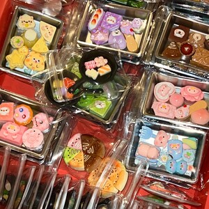 May include: A collection of miniature food-themed charms in clear plastic containers. The charms include sushi, pastries, and other cute food items in various colors. The containers are arranged in a red tray.