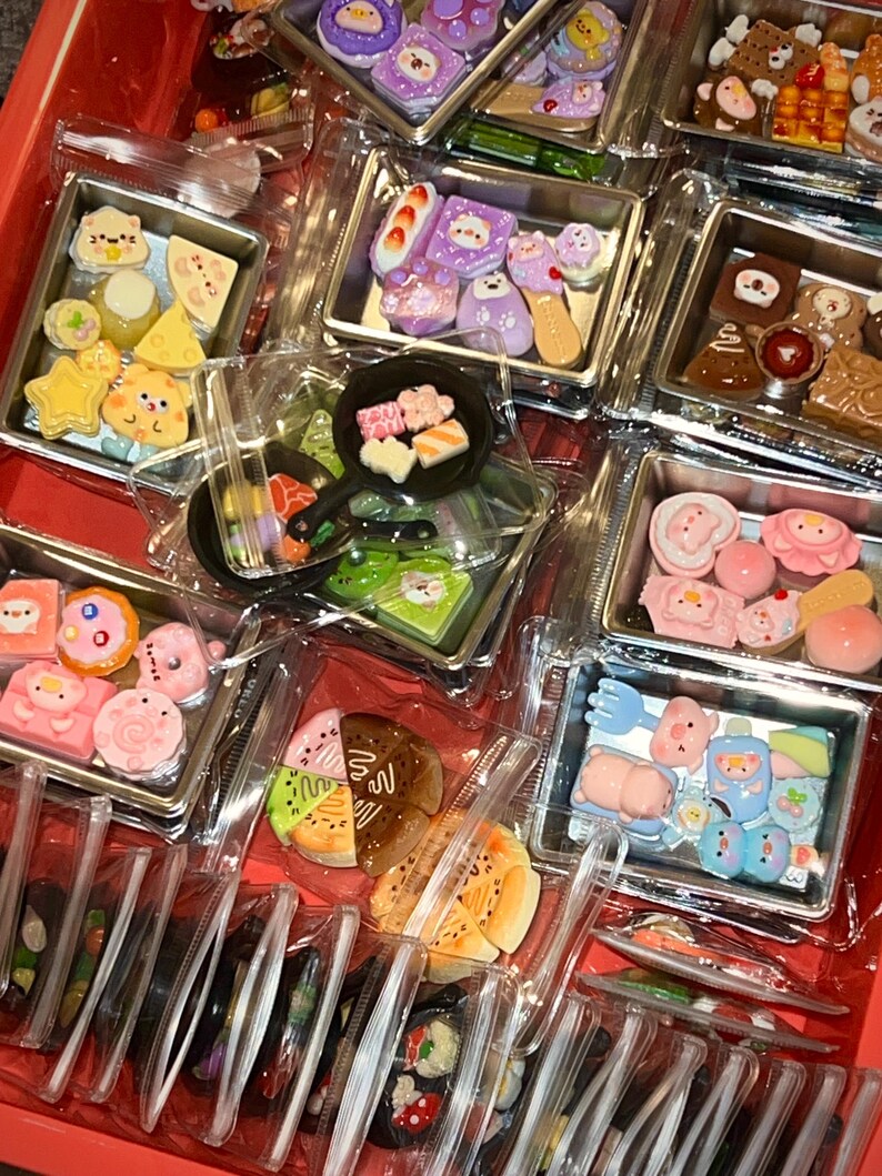 May include: A collection of miniature food-themed charms in clear plastic containers. The charms include sushi, pastries, and other cute food items in various colors. The containers are arranged in a red tray.