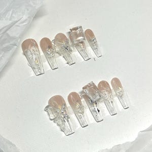 May include: A set of ten clear acrylic nails with a nude base. Each nail features intricate designs, including silver glitter, floral patterns, and 3D butterfly and cube accents. The nails are arranged on a white surface.